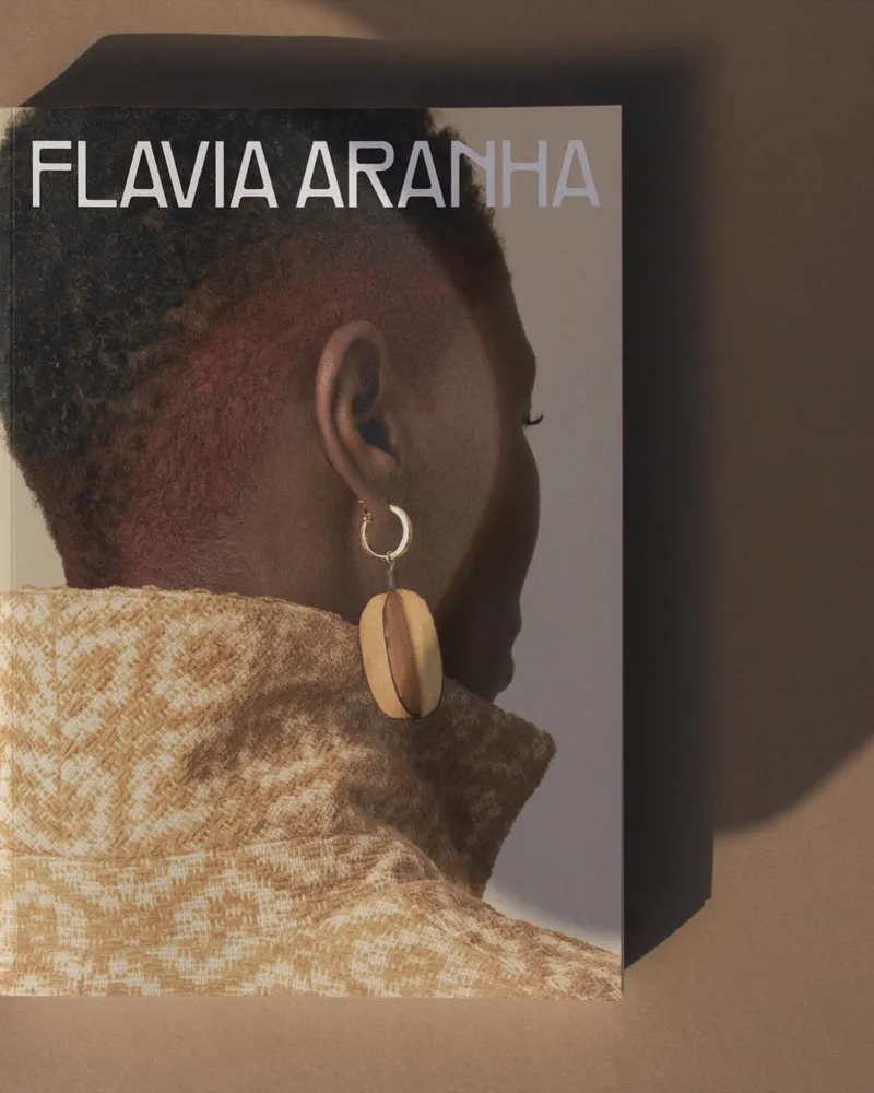 Lookbook da Flavia Aranha, marca do grupo Teia, sobre fundo areia: na capa, mulher de costas com brinco de madeira
